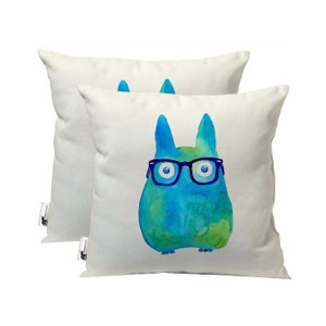 May include: Two white throw pillows with a blue and green watercolor illustration of a creature wearing glasses. The creature has large eyes and a round body.