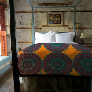 May include: A four-poster bed with a vibrant, patterned duvet cover in shades of orange, teal, and red. The bed is made up with white pillows and a neutral-colored bed skirt. A framed painting hangs on the textured wall above the bed.