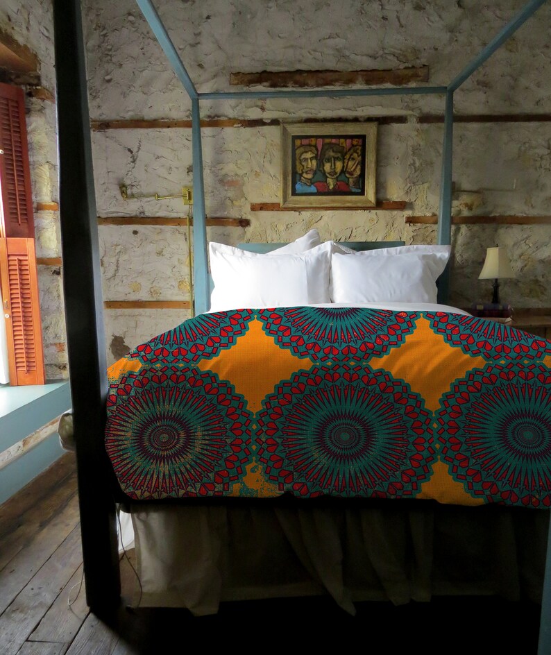 May include: A four-poster bed with a vibrant, patterned duvet cover in shades of orange, teal, and red. The bed is made up with white pillows and a neutral-colored bed skirt. A framed painting hangs on the textured wall above the bed.