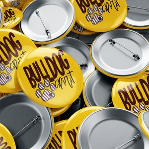 Puede incluir: Primer plano de un mont&oacute;n de botones redondos amarillos con el texto "Bulldog Spirit" y un dise&ntilde;o de huella de pata. Los botones tienen la parte posterior plateada con imperdibles.