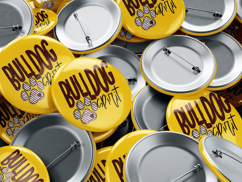 Puede incluir: Primer plano de un mont&oacute;n de botones redondos amarillos con el texto "Bulldog Spirit" y un dise&ntilde;o de huella de pata. Los botones tienen la parte posterior plateada con imperdibles.