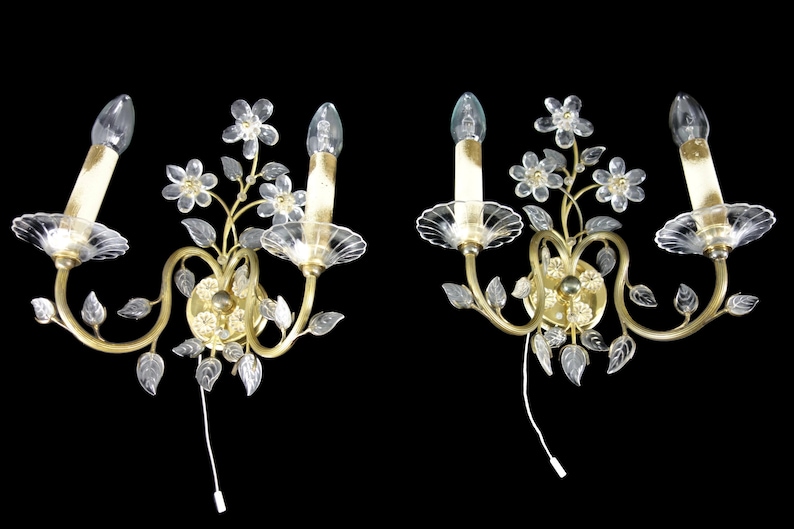 Pu&ograve; includere: Un paio di applique da parete dorate con fiori e foglie in vetro trasparente. Ogni applique ha due lampadine a forma di candela.