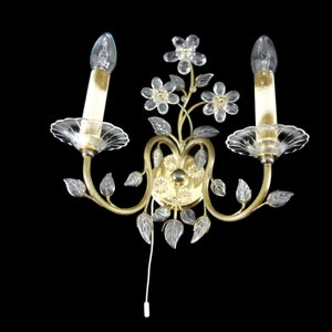 Pu&ograve; includere: Un'applique da parete dorata con due lampadine a candela in vetro trasparente e dettagli floreali e fogliari in cristallo.