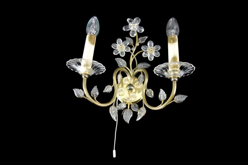 Pu&ograve; includere: Un'applique da parete dorata con due lampadine a candela in vetro trasparente e dettagli floreali e fogliari in cristallo.