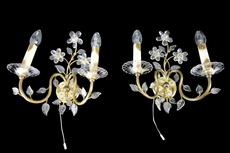 Pu&ograve; includere: Un paio di applique da parete dorate con fiori e foglie in vetro trasparente. Ogni applique ha due lampadine a forma di candela.