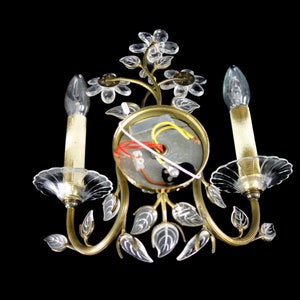 Pu&ograve; includere: Un'applique da parete dorata con due lampadine in vetro trasparente a forma di candela. L'applique &egrave; decorata con fiori e foglie in vetro trasparente.