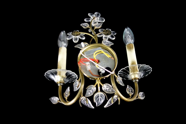 Pu&ograve; includere: Un'applique da parete dorata con due lampadine in vetro trasparente a forma di candela. L'applique &egrave; decorata con fiori e foglie in vetro trasparente.
