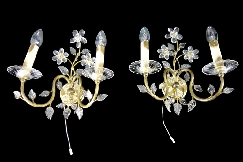 Pu&ograve; includere: Un paio di applique da parete dorate con fiori e foglie in vetro trasparente. Ogni applique ha due lampadine a forma di candela.