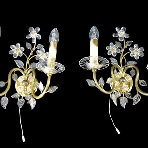 Pu&ograve; includere: Un paio di applique da parete dorate con accenti floreali in vetro trasparente e lampadine a forma di candela.