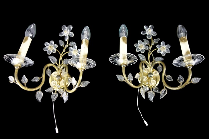 Pu&ograve; includere: Un paio di applique da parete dorate con accenti floreali in vetro trasparente e lampadine a forma di candela.