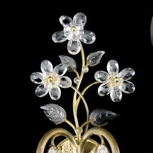 Pu&ograve; includere: Un'applique da parete dorata con due lampadine in vetro trasparente e un motivo floreale in cristallo.
