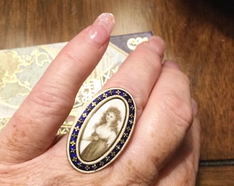 May include: An oval-shaped ring with a blue enamel border and a white enamel centre. The centre features a portrait of a woman in a white dress with brown hair. The ring is set with small gold accents.