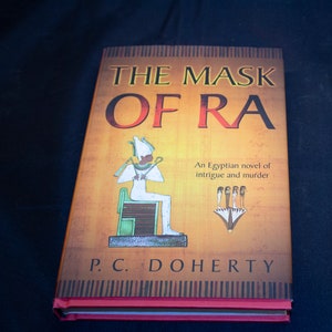 May include: A yellow paperback book with the title "The Mask of Ra" in large, bold letters. The book cover features an illustration of an Egyptian pharaoh sitting on a throne, with the text "An Egyptian novel of intrigue and murder" below the illustration. The author's name, "P.C. Doherty", is printed in large letters at the bottom of the cover.