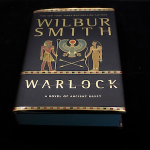 May include: A black hardcover book with gold lettering titled "Warlock" by Wilbur Smith. The book cover features an illustration of two figures in ancient Egyptian attire, a winged creature, and crossed arrows.