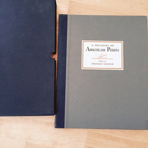 May include: A hardcover book titled "A Treasury of American Prints" with a gray cover and a rectangular label. The label features the title and the author's name, Thomas Craven. The book is next to a dark blue slipcase.