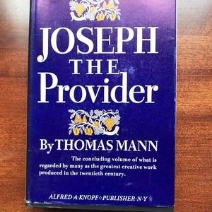 May include: A hardcover book titled "JOSEPH THE Provider" by Thomas Mann. The cover is a deep blue with white lettering and decorative illustrations. The book is a concluding volume of a creative work.