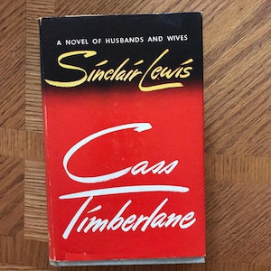 May include: A red hardcover book with the title "Cass Timberlane" in white cursive writing. The author, Sinclair Lewis, is written in yellow cursive writing above the title. The book cover also states "A Novel of Husbands and Wives".