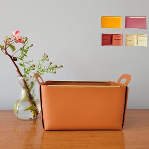 May include: A brown leather storage basket with gold accents and two handles. The basket is sitting on a wooden table with a vase of flowers in the background.