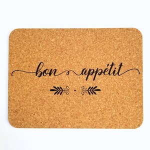 May include: A cork coaster with the text "bon appétit" in black cursive lettering and a decorative leaf design.