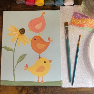 May include: A painting kit with a canvas featuring three colorful birds and a sunflower. The kit includes paint bottles in various colors, two paint brushes, and a palette. The text "Made by Kate" is visible on a small card.