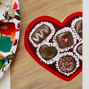 May include: A painting of a heart-shaped box of chocolates. The box is outlined in red and filled with various chocolate candies in white paper cups. The background is a light brown color. A paint palette is in the upper left corner.