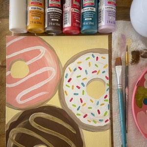 May include: A canvas painting of three donuts. The donuts are painted in a realistic style with different colors and toppings. The donuts are on a yellow background. The painting is surrounded by paint bottles and brushes.