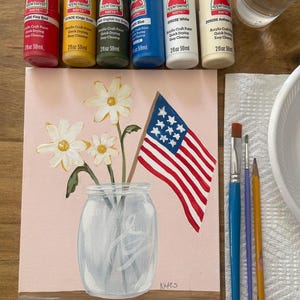 May include: A painting of a mason jar with white daisies and an American flag. The painting is on a pink background with a wooden base. Paint bottles in red, yellow, green, blue, and white are arranged above the painting.