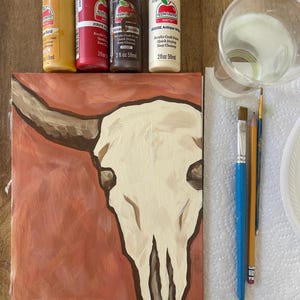 May include: A painted canvas featuring a cream-colored bull skull against a reddish-brown background. Four bottles of acrylic paint in gold, red, brown, and antique white are visible, along with paintbrushes and a cup of water. The painting is on a wooden surface.