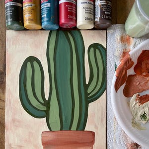 May include: A painted cactus in a terracotta pot on a light pink background. The cactus is painted in shades of green. Paint bottles in various colors are arranged above the painting, along with a palette of mixed paint colors.