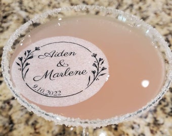 May include: A pink cocktail in a martini glass with a sugared rim. The glass has a sticker with the text "Aiden & Marlene 9.10.2022" in a circle with floral designs.