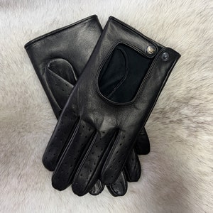 May include: A pair of black leather driving gloves featuring perforated detailing on the fingers and palm area, a cutout on the back of the hand, and a small snap button closure at the wrist. The gloves have a smooth finish and stitched seams.
