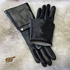 May include: A pair of black leather gloves with a zipper detail on the cuff. The gloves are folded in half and laid flat on a white fur surface.
