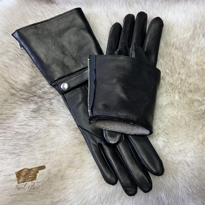 May include: A pair of black leather gloves with a zipper detail on the cuff. The gloves are folded in half and laid flat on a white fur surface.