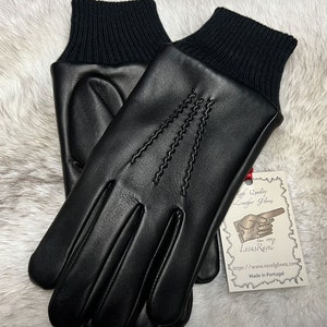 Puede incluir: Un par de guantes de cuero negro con puño de punto acanalado. Los guantes tienen un diseño cosido en el dorso de la mano y están hechos en Portugal. La etiqueta dice "High Quality Leather Gloves,  Lovas Ravel, Since 1999, https://www.ravelgloves.com, Made in Portugal."