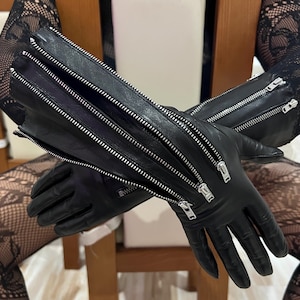 May include: A pair of black leather gloves featuring multiple silver-toned metal zippers running vertically along the forearm and wrist area. The gloves have a smooth finish and include several zipper pulls, with one zipper partially open.