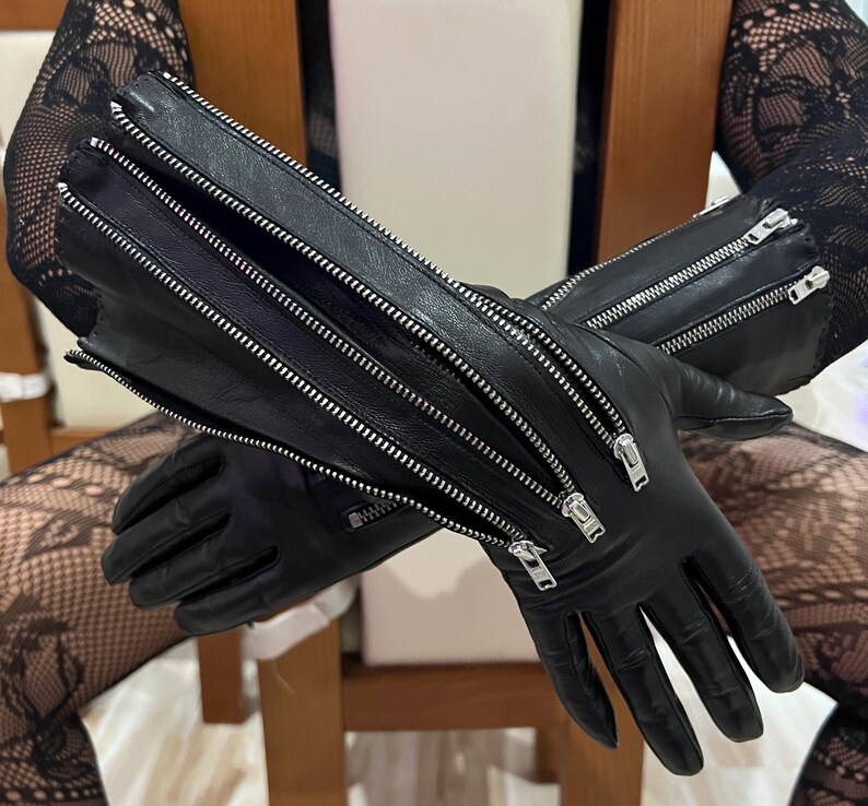 May include: A pair of black leather gloves featuring multiple silver-toned metal zippers running vertically along the forearm and wrist area. The gloves have a smooth finish and include several zipper pulls, with one zipper partially open.