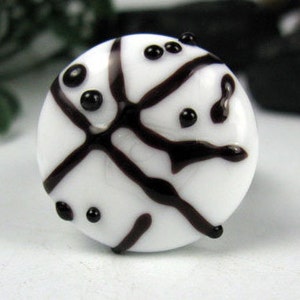 Könnte beinhalten: Ein weißer Glasring mit einem schwarzen, abstrakten Design. Der Ring hat eine runde, polierte Oberfläche mit dunkelbraunen Linien und kleinen schwarzen Punkten. Das Design erinnert an ein Netzwerk oder ein stilisiertes Muster.