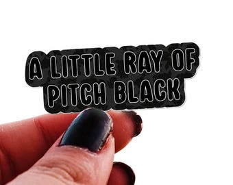 May include: A black sticker with the text "A LITTLE RAY OF PITCH BLACK" in a bold, outlined font. The sticker is held by a hand with dark nail polish against a white background.