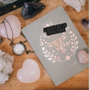 May include: A pale green journal featuring a rose gold Taurus design and the text "A LITTLE RAY OF PITCH BLACK." Pink heart-shaped stones and a pendant necklace are arranged on a wooden surface.