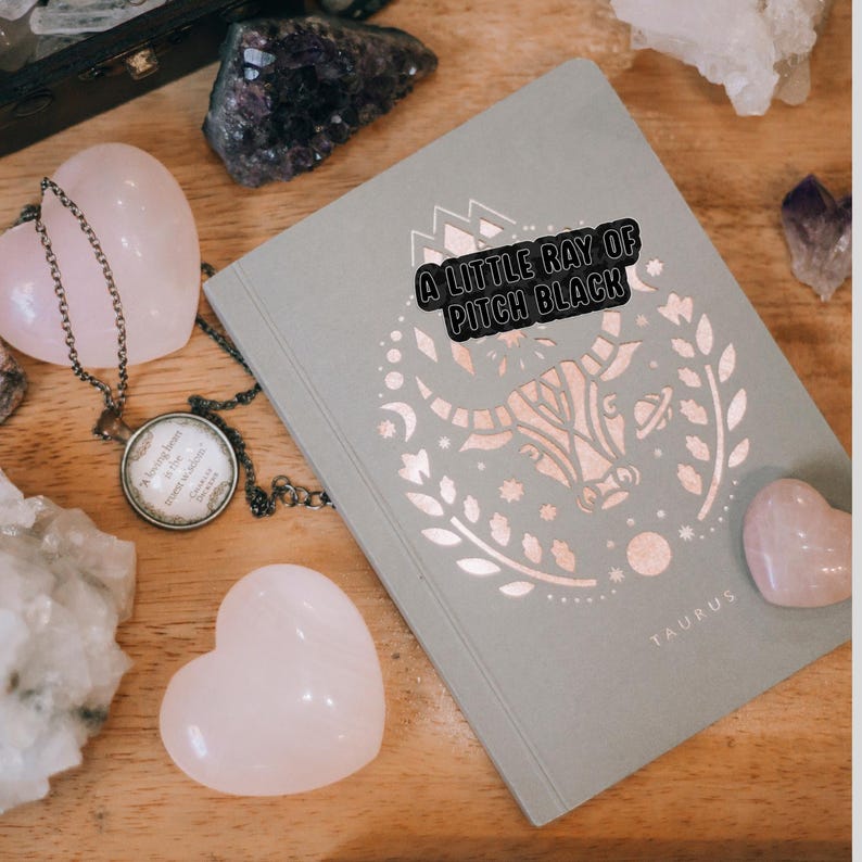 May include: A pale green journal featuring a rose gold Taurus design and the text "A LITTLE RAY OF PITCH BLACK." Pink heart-shaped stones and a pendant necklace are arranged on a wooden surface.