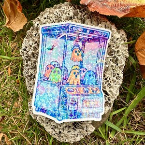 Könnte beinhalten: Ein Aufkleber mit einer blauen und violetten Glitzer-Greifmaschine mit dem Text "Grab A Ghost" und mehreren weißen Geistern darin.