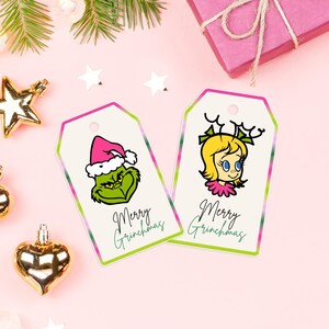 Könnte beinhalten: Zwei Weihnachtsgeschenkanhänger mit Grinch-Illustrationen. Ein Anhänger zeigt den Grinch mit Weihnachtsmütze, der andere ein Mädchen mit Geweih. Beide Anhänger tragen den Text "Merry Grinchmas" in grüner Schrift. Die Anhänger haben einen rosa-grünen Rand.
