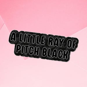 May include: A black sticker with white outline text that reads "A LITTLE RAY OF PITCH BLACK" on a pink background. The sticker has a floral pattern within the black area.