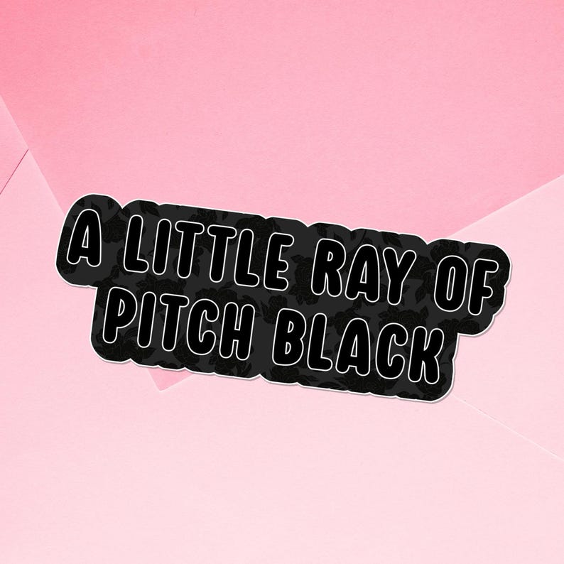 May include: A black sticker with white outline text that reads "A LITTLE RAY OF PITCH BLACK" on a pink background. The sticker has a floral pattern within the black area.