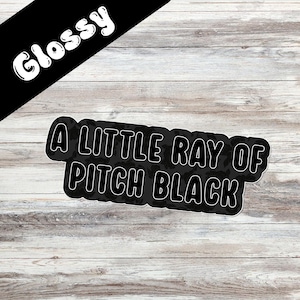 May include: A black sticker with the text "A LITTLE RAY OF PITCH BLACK" in white, outlined in black. The word "Glossy" is written in white on a black banner in the upper left corner. The sticker is on a light wood grain background.