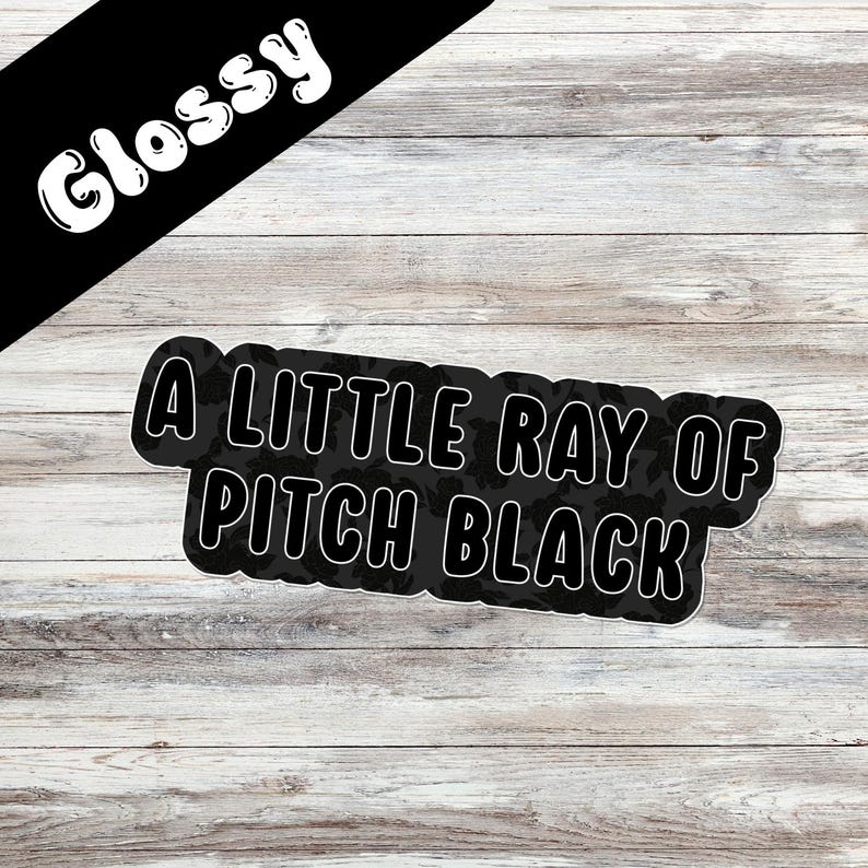 May include: A black sticker with the text "A LITTLE RAY OF PITCH BLACK" in white, outlined in black. The word "Glossy" is written in white on a black banner in the upper left corner. The sticker is on a light wood grain background.
