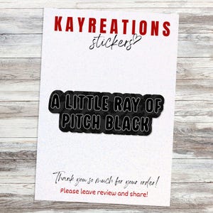 May include: A sticker featuring the phrase "A LITTLE RAY OF PITCH BLACK" in a black and white design. The sticker is on a white background with the words "KAYREATIONS stickers" in red. Below the sticker is the text "Thank you so much for your order! Please leave review and share!"