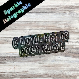 May include: A holographic sticker with the text "A LITTLE RAY OF PITCH BLACK" in white, outlined in black. The sticker has a black background with a rainbow sheen. The words "Sparkle Holographic" are in the upper left corner.