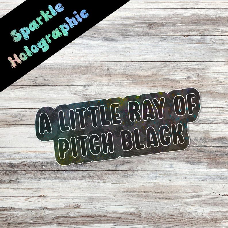 May include: A holographic sticker with the text "A LITTLE RAY OF PITCH BLACK" in white, outlined in black. The sticker has a black background with a rainbow sheen. The words "Sparkle Holographic" are in the upper left corner.