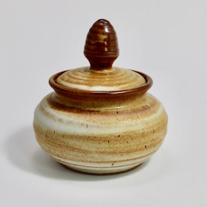 May include: A small, round, ceramic jar with a lid. The jar is decorated with brown and white stripes and has a brown knob on the lid.